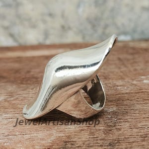 Könnte beinhalten: Ein polierter Silberring mit einem fließenden, asymmetrischen Design. Der Ring hat eine breite, geschwungene Oberseite, die sich zu einer Spitze verjüngt, und ein Band, das den Finger umschließt. Der Ring liegt auf einer Holzoberfläche.