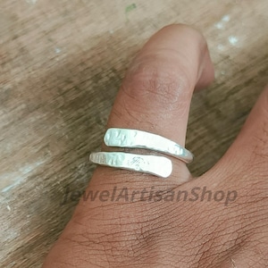 May include: A silver ring with a hammered texture, wrapping around a finger. The ring has two overlapping bands. The background is a wooden surface. The text "JewelArtisanShop" is visible.