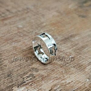 May include: A silver ring with a world map design. The ring is made of shiny metal and features cut-out shapes of continents and islands. The ring is displayed on a wooden surface.