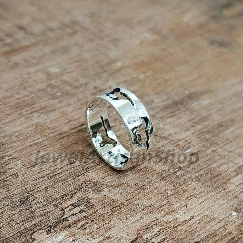 May include: A silver ring with a world map design. The ring is made of shiny metal and features cut-out shapes of continents and islands. The ring is displayed on a wooden surface.