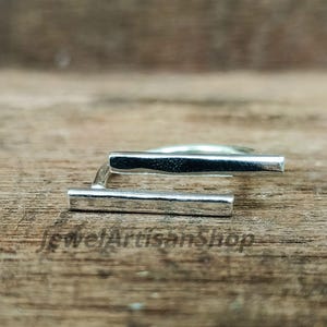 Könnte beinhalten: Ein silberner Ring mit modernem Design. Der Ring hat zwei rechteckige Stäbe, die in einem rechten Winkel verbunden sind. Der Ring ist vor einem Holzgrund platziert.