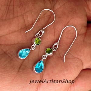 Könnte beinhalten: Silberne Ohrringe mit einem tropfenförmigen, hellblauen Edelstein und einem runden, grünen Edelstein. Die Ohrringe haben ein Haken-Design. Der Text "JewelArtisanShop" ist sichtbar.