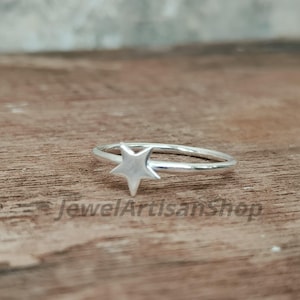 May include: A silver ring with a star-shaped design. The ring is set against a wooden surface. The text "JewelArtisanShop" is visible in the image. This ring is a piece of jewelry.