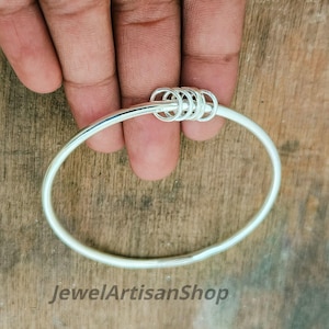 Könnte beinhalten: Ein silbernes Armreif-Armband mit einer Reihe kleiner, verbundener Ringe oben. Das Armband wird von einer Hand auf einer Holzoberfläche gehalten. Der Text "JewelArtisanShop" ist unten im Bild zu sehen.