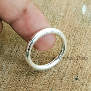Peut inclure: Une bague en argent poli avec un anneau arrondi. La bague est tenue entre deux doigts sur une surface en bois. La surface lisse de la bague reflète la lumière, soulignant sa forme circulaire.