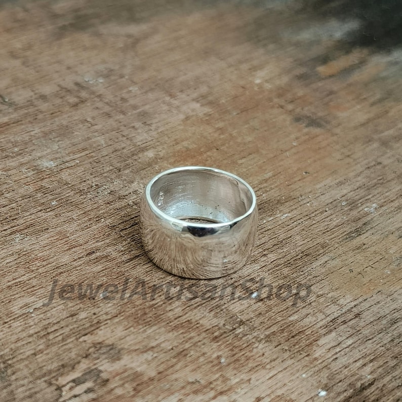 Op de afbeelding: Een brede, gepolijste zilveren ring met een glad, afgerond ontwerp. De ring is geplaatst op een houten oppervlak met textuur. De eenvoudige, maar elegante stijl van de ring maakt het een veelzijdig accessoire.