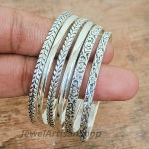 Könnte beinhalten: Eine Sammlung silberner Armreifen mit aufwendigen Designs. Die Armreifen weisen Muster wie Blätter und florale Motive auf. Der Schmuck wird auf einer Hand präsentiert, die die Details der Metallarbeiten zeigt.