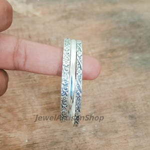 May include: Three silver rings are stacked together, with a floral pattern on the outer rings and a smooth, polished band in the centre. The rings are held by a finger against a wooden background. The text "JewelArtisanShop" is visible.