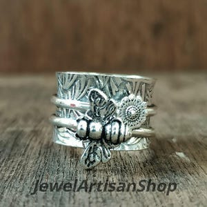 Pode incluir: Um anel giratório prateado com um design de abelha e flor. O anel apresenta uma abelha detalhada e uma pequena flor na banda. O anel tem um padrão floral texturizado e o texto "JewelArtisanShop".