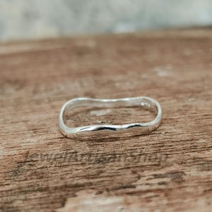 Può includere: Un anello in argento sterling con un design ondulato unico. L'anello ha una finitura liscia e lucida e una forma leggermente irregolare. L'anello è mostrato su una superficie di legno.