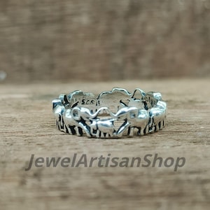 May include: A silver ring with a repeating elephant design. The ring has a detailed, openwork pattern with multiple elephants. The text "925" is visible on the ring. The ring is on a wooden surface. The shop name "JewelArtisanShop" is visible.
