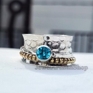 Könnte beinhalten: Ein silberner Spinner-Ring mit gehämmertem Muster und einem runden, leuchtend blauen Edelstein. Der Ring hat ein goldfarbenes Band und den Text "JewelArtisanShop".
