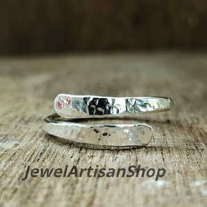 Könnte beinhalten: Ein silberfarbener Wickelring mit gehammerter Textur. Der Ring hat ein offenes Design, wobei sich die Enden überlappen. Der Ring wird auf einer Holzoberfläche präsentiert. Der Text "JewelArtisanShop" ist unten im Bild zu sehen.