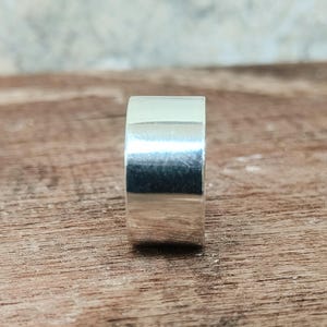 May include: A close-up of a silver ring with a flat, wide band. The ring is highly polished and reflects light, appearing bright against a wooden surface. The ring's design is simple and modern, suitable for everyday wear.