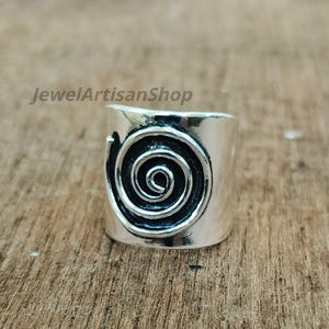 Könnte beinhalten: Ein breiter, silberfarbener Ring mit einem markanten Spiraldesign. Der Ring hat eine zylindrische Form mit poliertem Finish, und die Spirale ist mittig auf der Ringoberfläche platziert. Der Text "JewelArtisanShop" ist sichtbar.