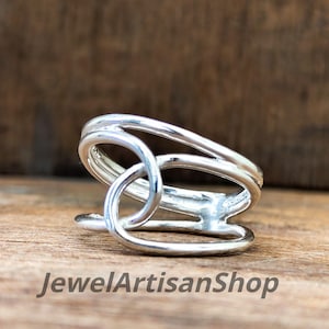 May include: A silver-colored ring with an open, geometric design. The ring features a series of curved bands and an oval-shaped element. The text "JewelArtisanShop" is visible at the bottom of the image. The ring is displayed on a wooden surface.