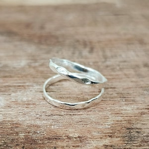 May include: Two silver rings with a textured, hammered finish are stacked on a wooden surface. The rings have an organic, irregular shape, reflecting light and creating highlights. The rings are likely jewelry.