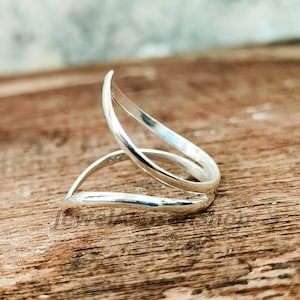 Peut inclure: Une bague en argent au design ouvert unique. La bague présente deux bandes incurvées qui se chevauchent, créant un look élégant et moderne. Le métal poli reflète la lumière, soulignant la surface lisse de la bague. La bague est présentée sur une surface en bois.