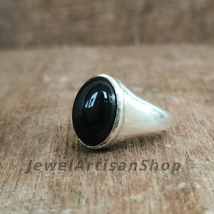 May include: A silver ring with a large, oval, black onyx stone. The ring has a polished finish and is set on a wooden surface. The text "JewelArtisanShop" is visible on the surface.