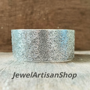 Könnte beinhalten: Ein breites silbernes Armreif-Armband mit einem aufwendigen Blumenmuster. Das Armband besteht aus glänzendem Metall und hat ein detailliertes, geprägtes Design. Der Text "JewelArtisanShop" befindet sich unten.