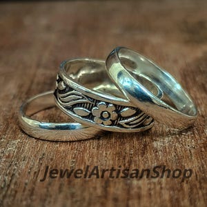 Puede incluir: Tres anillos de plata sobre una superficie de madera. Un anillo presenta un diseño floral, mientras que los otros dos son bandas lisas. Los anillos se muestran con el texto "JewelArtisanShop" en la parte inferior de la imagen.