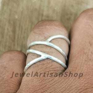 May include: A silver ring with an overlapping X design. The ring is worn on a finger. The text "JewelArtisanShop" is visible in the image.