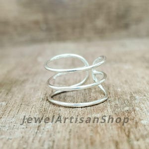 May include: A silver-colored ring with a unique, open design. The ring features three spiraling bands that wrap around the finger. The ring is displayed on a wooden surface. The text "JewelArtisanShop" is visible at the bottom.