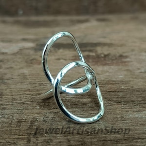 Könnte beinhalten: Ein silberner Ring mit abstraktem Design. Der Ring hat zwei ineinander verschlungene ovale Formen, die einen einzigartigen und modernen Look erzeugen. Der Ring ist auf einer Holzoberfläche platziert, was seine polierte Oberfläche hervorhebt.