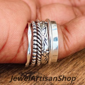 Könnte beinhalten: Eine Ansammlung von Silberringen mit unterschiedlichen Designs, darunter ein glattes Band, ein Perlenband, ein gedrehtes Seilband und ein floral graviertes Band. Die Ringe sind auf einem Finger gestapelt und zeigen die verschiedenen Texturen. Der Text "Jewel Artisan Shop" ist sichtbar.
