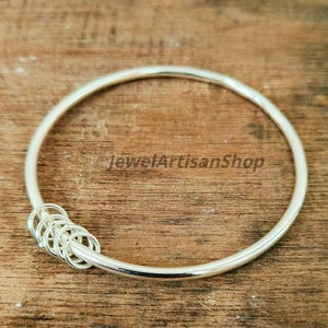Könnte beinhalten: Ein silbernes Armreif-Armband mit einer Reihe kleiner, miteinander verbundener Ringe auf einer Seite. Das Armband hat eine glatte, polierte Oberfläche und wird auf einer Holzoberfläche präsentiert. Der Text "JewelArtisanShop" ist sichtbar.