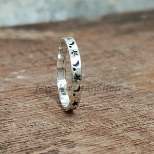Puede incluir: Un anillo de plata con un patrón repetido de estrellas y lunas crecientes. El anillo es delgado y tiene un acabado pulido. El diseño celestial está grabado en la banda, creando un aspecto delicado y detallado. El anillo se muestra sobre una superficie de madera.