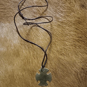 May include: A brass-colored pendant in the shape of a cross hangs from a dark brown leather cord. The pendant has a circular hole at the top for the cord. The background is a textured, brown surface.