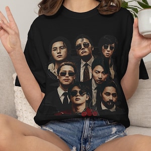Peut inclure: Un t-shirt noir à manches courtes arborant une impression graphique de sept personnes portant des costumes et des lunettes de soleil. Le motif est disposé dans un style collage avec une esthétique sombre et mystérieuse.