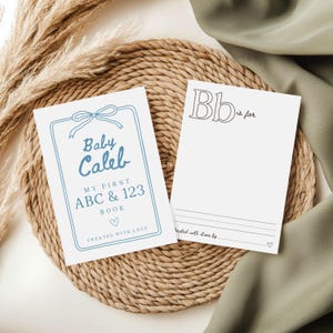 Puede incluir: Una portada de libro para bebé azul y blanco con el texto "Baby Caleb" y "My First ABC & 123 Book" con un símbolo de corazón. La portada está sobre una estera de paja tejida con un fondo blanco.