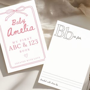 Könnte beinhalten: Ein rosa-weißer Babybuchumschlag mit Schleife und dem Text "Baby Amelia My First ABC & 123 Book Created with Love".