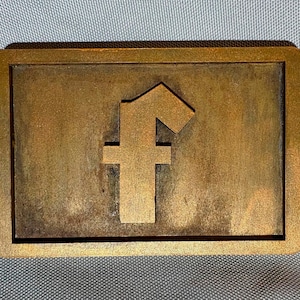 May include: A rectangular, bronze-colored belt buckle featuring a raised letter "f" in the center. The buckle has a slightly weathered appearance with a darker border and a lighter, textured center. The edges are rounded.