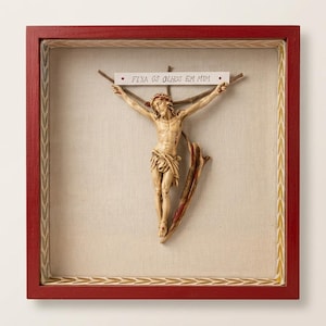 May include: A framed religious artwork featuring a carved figure of Jesus on a cross. The figure is beige with red accents, mounted on a beige fabric background within a red frame. A small sign above reads "FIXA OS OLHOS EM MIM".