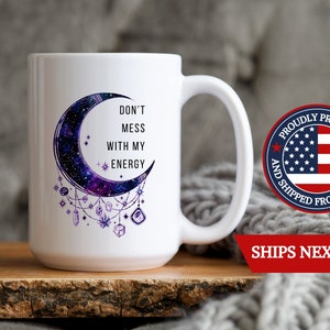 May include: White ceramic mug with a crescent moon design featuring a galaxy print and purple crystals hanging from it. The text "Don't Mess With My Energy" is printed on the mug.