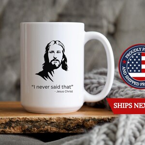 May include: White ceramic mug featuring a black silhouette of Jesus with the quote "I never said that" and the name "Jesus Christ." The mug has a large handle and is displayed on a wooden surface.