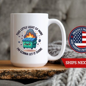 May include: White ceramic coffee mug with a green trash can with a smiling face and flames coming out of it. The text on the mug reads "This little light of mine I'm gonna let it shine".
