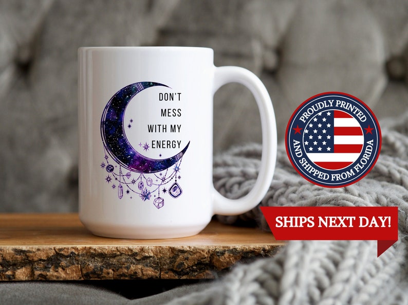 May include: White ceramic mug with a crescent moon design featuring a galaxy print and purple crystals hanging from it. The text "Don't Mess With My Energy" is printed on the mug.