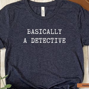 May include: A dark heather navy blue t-shirt with the text "BASICALLY A DETECTIVE" printed in white.