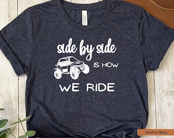 May include: Heather navy t-shirt with white text and a graphic of an off-road vehicle. The text reads "side by side is how we ride". The shirt has a crew neck and short sleeves.
