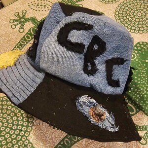 May include: A blue denim bucket hat with a black brim and a black fabric patch on the brim. The hat has a black fabric patch on the crown with the letters "CP" written in black.