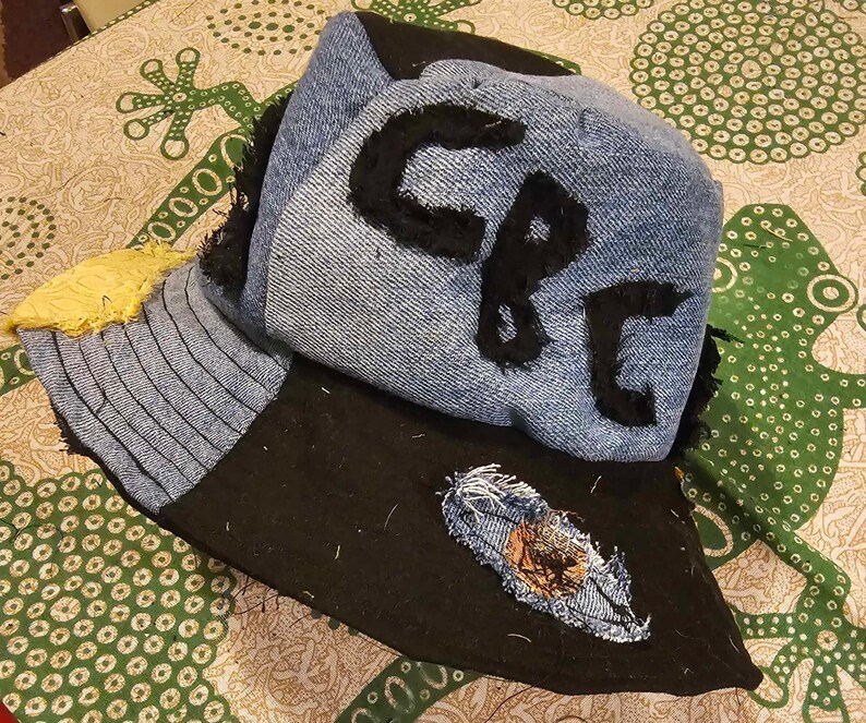 May include: A blue denim bucket hat with a black brim and a black fabric patch on the brim. The hat has a black fabric patch on the crown with the letters "CP" written in black.