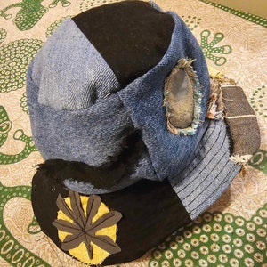 以下が含まれることがあります： 青いデニムの帽子で、黒い布のクラウンと黄色の葉のパッチが付いています。帽子は、縁がほつれていて、側面に穴が開いているなど、使い古したような外観をしています。
