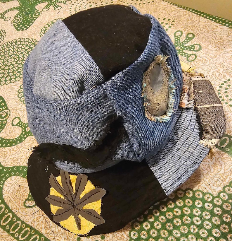 May include: A blue denim hat with a black fabric crown and a yellow leaf patch. The hat has a distressed look with frayed edges and a hole in the side.