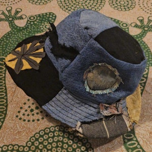 May include: A blue, black, and yellow patchwork hat with a circular window. The hat is made of different fabrics and has a unique, handmade style.