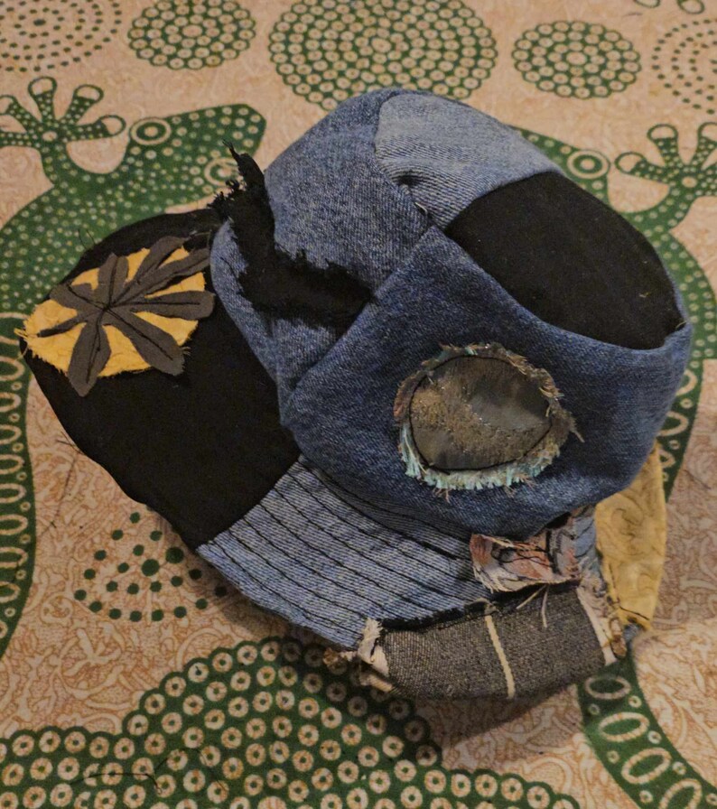 May include: A blue, black, and yellow patchwork hat with a circular window. The hat is made of different fabrics and has a unique, handmade style.