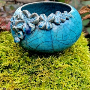 May include: A small, teal blue ceramic bowl with a crackled glaze and raised flower details around the rim. The bowl is sitting on a bed of green moss.